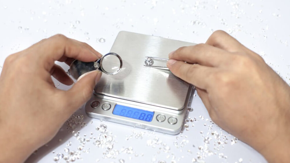 Hand holding a diamond with weight scale tweezers and a pile of diamond